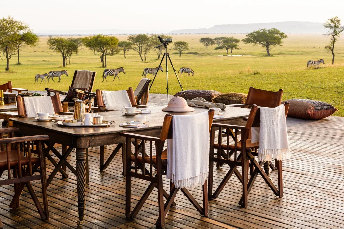 A breakfast table set on the deck of Singita Sabora Tented Camp with zebra on the plain beyond, the luxury end of Serengeti safari packages