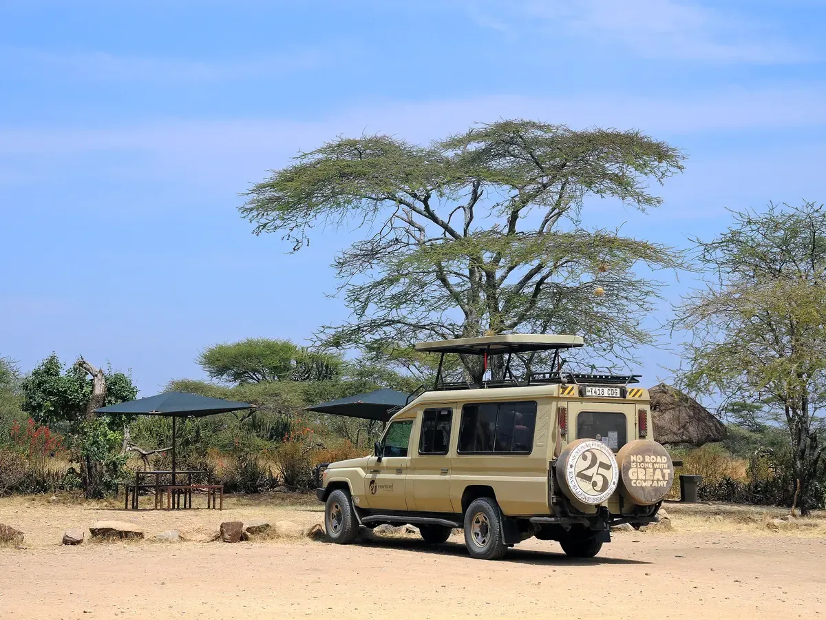 A safari Land Cruiser with a pop-up roof parked under an acacia at a camp stop in Tanzania, typical of Serengeti safari packages